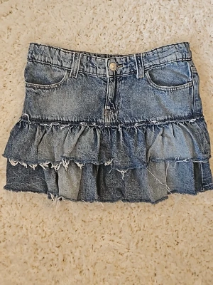 Blå jeanskjol med volanger - Cool kort jeanskjol i blå denim med två lager volanger och råa, fransiga kanter. Kjolen har klassiska fickor fram och bak samt bälteshällor. Perfekt för dig som gillar en trendig och avslappnad stil.