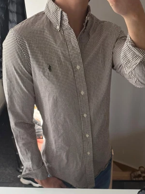 Rutig skjorta från Ralph Lauren - Klassisk rutig skjorta från Ralph Lauren i brunt och vitt. Skjortan har lång ärm, knappar framtill och button-down krage. Tillverkad i mjuk bomull och har den ikoniska broderade loggan på bröstet. Perfekt för en stilren och avslappnad look.