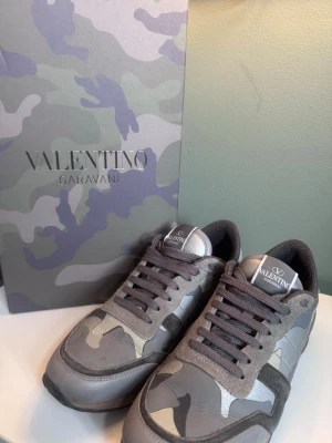 Valentino Garavani camo sneakers grå - Snygga sneakers från Valentino Garavani i grått camouflagemönster. Skorna har grå mesh och mockadetaljer, samt klassisk snörning och logga på plösen. Perfekta för dig som vill sticka ut med stilrena och exklusiva sneakers.