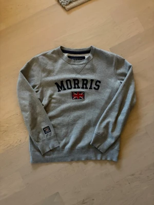 Grå sweatshirt från Morris (beskrivning) - Snygg grå sweatshirt från Morris med broderad logga och brittisk flagga på bröstet. Tröjan har rund halsringning, ribbade muddar och en klassisk passform. Perfekt för en avslappnad och stilren look. Skickar helst med instabox! 