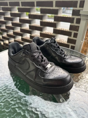 Svarta Nike Air Force 1 sneakers - Säljer ett par klassiska helsvarta Nike Air Force 1 sneakers i läder. Skorna har lågt skaft, perforerad tåbox och ikonisk swoosh på sidorna. Tjock sula och metall-detalj på snörningen med 'AF1'. Perfekta för en clean och stilren look.