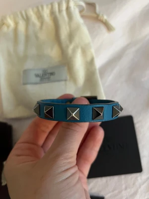 Blått Valentino Armband med nitar i läder - Säljer detta blåa valentino armband i läder, köpbevis och låda medföljer. Har också ett gammalt kvitto från onlinebeställning. Armbandet har spruckit lite, syns på näst sista bilden, men ser väldigt bra ut på. Annars i fint skick. Medföljer en extra stor nit.