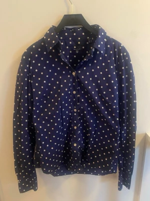 Blå prickig skjorta från GANT - Snygg blå skjorta från GANT med vita prickar och klassisk krage. Skjortan har långa ärmar och knäppning framtill. 