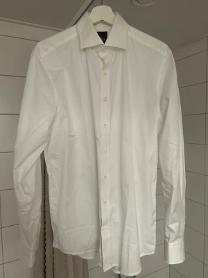 Vit klassisk skjorta med knappar - Säljer en stilren vit skjorta med klassisk krage och långa ärmar. Skjortan har knäppning framtill och är tillverkad i ett slätt bomullstyg. Perfekt för dig som gillar en clean och tidlös look.