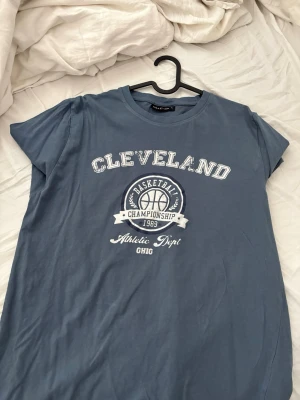 Blå Cleveland t-shirt från Reaction - Snygg blå t-shirt från Reaction med tryck 'Cleveland Basketball Championship 1983' på bröstet. Klassisk rund hals och korta ärmar. Perfekt för en sportig och avslappnad stil.