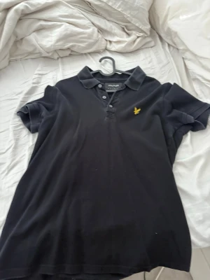 Svart pikétröja från Lyle & Scott - Klassisk svart pikétröja från Lyle & Scott med gul broderad logga på bröstet. Tröjan har korta ärmar, krage och knappar framtill. Perfekt för en stilren och avslappnad look.