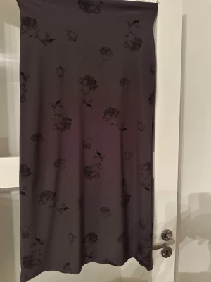  lång kjol - Säljer en lång, mörk kjol med  blommönster. Kjolen har en rak och enkel siluett och är tillverkad i ett mjukt, tunt material som faller snyggt. Perfekt för dig som gillar stilrena plagg med en twist. Nopprig nedantill i övrigt inget att anmärka på :)