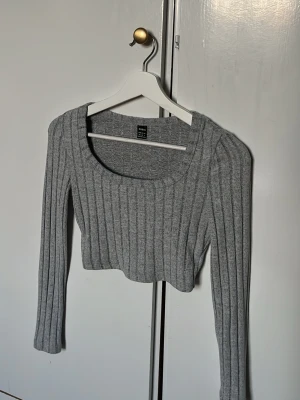 Grå ribbad cropped stickad tröja - Säljer en grå ribbstickad cropped tröja med lång ärm och bred, rundad urringning. Tröjan är en tight passform som framhäver siluetten. Perfekt att styla med höga jeans eller kjol för en trendig look. Magtröja typ 💗😍
