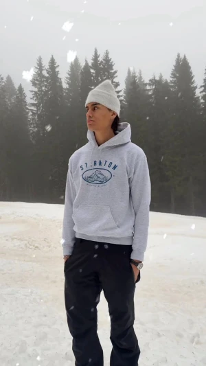 Grå hoodie St. Anton med tryck - Grå hoodie med blått St. Anton-tryck och illustration av berg på bröstet. Klassisk modell med huva och känguruficka framtill. Perfekt för chill dagar eller lager på lager-stil. Mjuk bomullskänsla och ribbade muddar vid ärmslut och nederkant. Helt ny, helt oanvänd