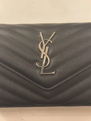 Svart YSL axelväska i läder - Snygg svart axelväska från YSL i strukturerat läder med silverfärgad logga framtill. Väskan har justerbar axelrem och klassisk kuvertformad design med chevronmönster. Perfekt för dig som vill ha en stilren och lyxig accessoar.