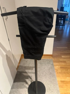 Svarta cargobyxor från Kappahl - KappAhl byxor för barn, unisex, i storlek 146. Byxorna är svarta och tillverkade i bomull och elestan. Modellen har knapp och dragkedja framtill, hällor för skärp samt flera fickor, inklusive benfickor. Passar vardagsbruk och fritidsaktiviteter för barn i storlek 146. Endast använda vid ett tillfälle. 