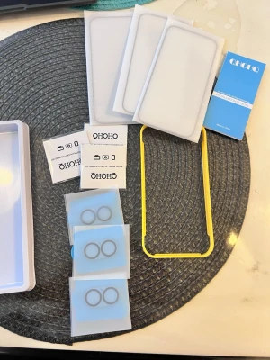 QHOHQ skärmskydd till iPhone 16 - Säljer ett paket QHOHQ härdat glas skärmskydd till iPhone 16. Innehåller tre genomskinliga skärmskydd, monteringsram i gul plast, rengöringsdukar och linsskydd till kameran. Skydden är tunna och har hög transparens för optimal touchkänsla.