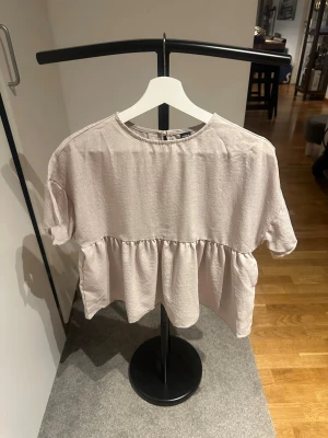 Ljusbeige volangblus med kort ärm - Supersöt ljusbeige blus med volangdetalj och korta ärmar. Blusen har en avslappnad passform och rund halsringning, perfekt för en chill och trendig look. 