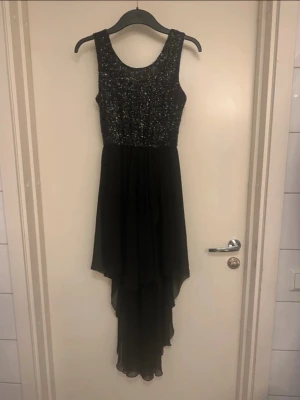 Svart glittrig långklänning Sisters Point  - Svart långklänning från Sisters Point med glittrig paljettöverdel och ärmlös design. Kjolen är i tunt, flygigt tyg med asymmetrisk längd som ger en snygg siluett. Perfekt för speciella tillfällen när du vill sticka ut.