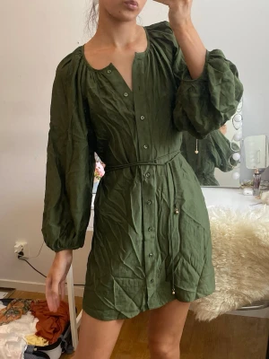Grön kort klänning från H&M - Stilren grön klänning från H&M i kort modell med långa puffärmar och knappar framtill. Klänningen har ett smalt knytband i midjan och är tillverkad i ett lätt och luftigt linnematerial. Perfekt för dig som gillar en avslappnad men trendig look.