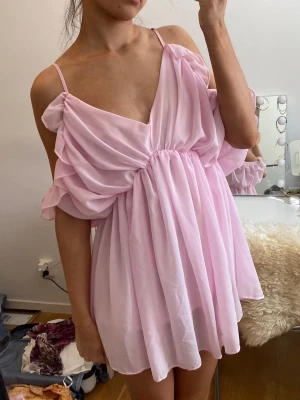 Ljusrosa off shoulder klänning - Supersöt ljusrosa klänning med off shoulder-design och tunna axelband. Klänningen har ett luftigt, flowy tyg och djup v-ringning. Voluminösa ärmar och rynkad midja ger en romantisk vibe. Perfekt för sommardagar eller när du vill känna dig extra söt.