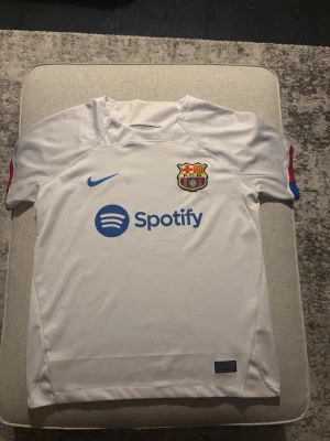 Vit FC Barcelona matchtröja Nike - Säljer en vit FC Barcelona matchtröja från Nike med klubbmärke och Spotify-logga på bröstet. Tröjan har korta ärmar, blå Nike-logga och detaljer i rött och blått på ärmarna. Tillverkad i lätt funktionsmaterial, perfekt för fotbollsträning eller match. Taggen i nacken har tvättats bort men är i storlek M