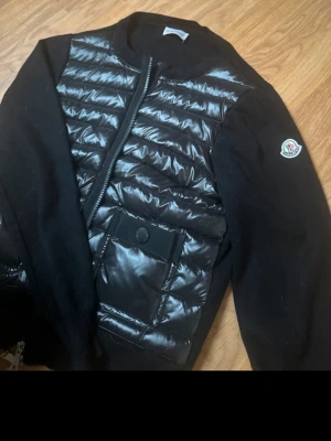Svart cardiganjacka från Moncler - Snygg svart cardiganjacka från Moncler med quiltad, glansig framsida och stickade ärmar. Jackan har två fickor framtill med knappar och dragkedja. Moncler-logga på ena ärmen. Perfekt för lager-på-lager och streetstyle. Pris kan diskuteras 