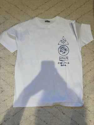Chrstian Dor - Vit t-shirt från Chrstian Dor med blå handritade tryck framtill och ett större motiv på ryggen. Klassisk passform med rund hals och korta ärmar. Materialet känns mjukt och skönt, perfekt för en stilren look.
