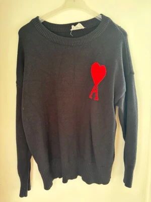 Svart stickad tröja Ami Paris - Säljer en svart stickad tröja från Ami Paris med deras ikoniska röda hjärta och bokstaven A broderat på bröstet. Tröjan har rund halsringning, långa ärmar och ribbade muddar. Perfekt för dig som gillar stilrena plagg med en twist.