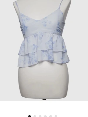 Ljusblå blommig volangtop - Supersöt ljusblå topp med blommigt mönster och dubbla volanger. Tunna justerbara axelband och v-ringad halsringning ger en feminin vibe. Perfekt för varma dagar och somriga outfits.