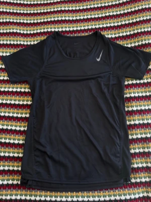 Svart Nike Tränings T-shirt - Svart kortärmad träningströja från Nike med Dri-FIT-teknologi som transporterar bort fukt. Tröjan har rund halsringning och en diskret Nike-logga på bröstet. Perfekt för löpning, gym eller annan sport.