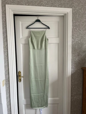 Ljusgrön långklänning med smala axelband - Snygg ljusgrön långklänning med smala axelband och rak halsringning. Klänningen har en minimalistisk design och är tillverkad i ett glansigt material som ger en elegant känsla. Perfekt för dig som gillar stilrena och enkla plagg. Aldrig använd, nypris 1 800kr