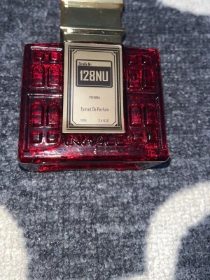 Inhale Vienna Extrait De Parfum 100ml - Lyxig parfym från Inhale, Vienna Extrait De Parfum, 100 ml. Flaskan är djupt röd med gulddetaljer och har en elegant, fyrkantig design med mönstrad yta. Kommer i en exklusiv guldig förpackning med vapensköld och text i svart och guld.
