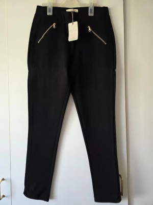 MQ svarta leggings med dragkedjor fuskfickor - Svarta leggings med smal passform och två snygga dragkedjor framtill, fuskfickor. Skön midja och stretchigt material som sitter bekvämt. Snygg avrundning vid byxslut. Perfekta till en avslappnad eller trendig look.