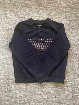 Marinblå sweatshirt från Emporio Armani - Stilren mörkblå sweatshirt från Emporio Armani med det ikoniska örnlogotypen i färgglatt mönster på bröstet. Tröjan har rund halsringning, ribbade muddar och är tillverkad i mjukt bomullsmaterial. Perfekt för en trendig och avslappnad look.