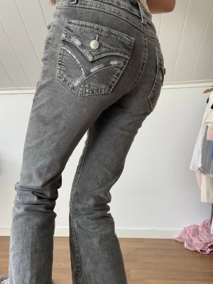 Grå bootcut jeans med broderade fickor - Snygga grå bootcut jeans med broderade detaljer och silvriga knappar på bakfickorna. Jeansen har en fin design på bakfickorna. 