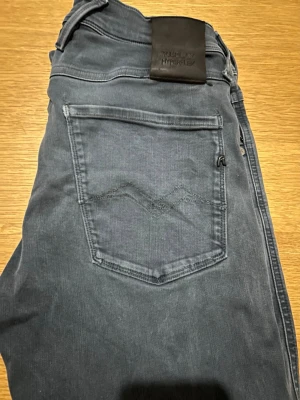 Mörkgrå jeans från Replay - Säljer ett par mörkgrå jeans från Replay med klassisk femficksdesign och snygga sömdetaljer på bakfickan. Jeansen har normal midja, smal passform och är tillverkade i mjukt denimtyg med stretch för extra komfort. Perfekta för en avslappnad och trendig look.