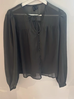 Olivgrön blus från Vero Moda - Snygg olivgrön blus från Vero Moda i tunn, transparent kvalitet. Blusen har långa ärmar, knappar framtill och en söt knytning vid halsen. Perfekt för dig som gillar en stilren och trendig look.