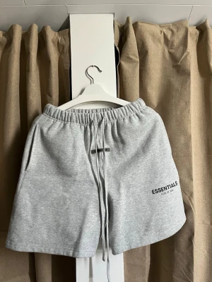 Essentialls Fear Of God Shorts - Säljer ett par klassiska Essentials Fear of God shorts i en snygg ljusgråmelerad färg. Shortsen är i väldigt bra skick och sparsamt använda. Detaljer: • Märke: Essentials Fear of God. • Storlek: Small (S). • Material: Mjuk och kraftig bomullsblandning (80% bomull, 20% polyester) med ruggad insida för maximal komfort. • Design: • Elastisk midja med extra långa dragskor. • Gummilogga framtill vid midjan samt tryckt logga på vänster ben. • Praktiska sidofickor samt en bakficka på höger sida.