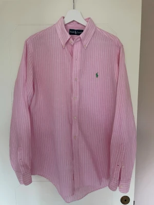 Rosa randig linneskjorta från Ralph Lauren - Grymt fin rosa skjorta från Ralph Lauren med vita ränder och broderad grön logga på bröstet. Tillverkad i ett lätt och svalt linnematerial, perfekt för en stilren look i sommar!