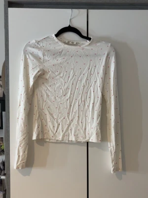 Vit långärmad topp med rosa hjärtan - Supersöt vit långärmad topp från 157 med små rosa hjärtan över hela tyget. Tillverkad i mjuk bomull och har en klassisk rund halsringning. Perfekt för dig som gillar gulliga detaljer och en enkel, clean stil.