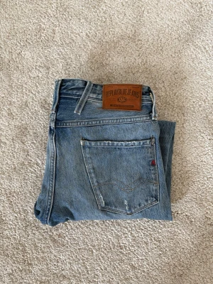 Replay blå slitna jeans - Säljer ett par blå jeans från Replay med snygga slitningar och klassisk femficksmodell. Jeansen har en läderpatch bak i midjan och detaljerade sömmar på bakfickan. Perfekta för en avslappnad och trendig look. På bild ser man den ända defekten på byxorna skriv vid funderingar 