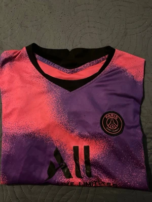 PSG matchtröja rosa/lila - Säljer en snygg PSG matchtröja i rosa och lila med svart V-ringning och klubbmärke på bröstet. Tröjan har trycket 'Accor Live Limitless' och är gjord i ett lätt och ventilerande material, perfekt för fotboll eller träning.
