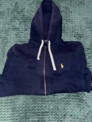 Marinblå hoodie från Polo Ralph Lauren - Snygg marinblå hoodie från Polo Ralph Lauren med dragkedja, vit dragsko och klassisk gul logga på bröstet. Tillverkad i mjuk bomullsblandning, perfekt för en avslappnad och stilren look. Huva och ribbade muddar ger extra komfort.