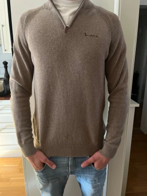 Beige tröja med halvzip från Bondelid - Snygg beige stickad tröja från Bondelid med halvzip och diskret broderad logga på bröstet. Tröjan har långa ärmar, ribbade muddar och en klassisk passform. Perfekt att bära över en t-shirt för en avslappnad stil.