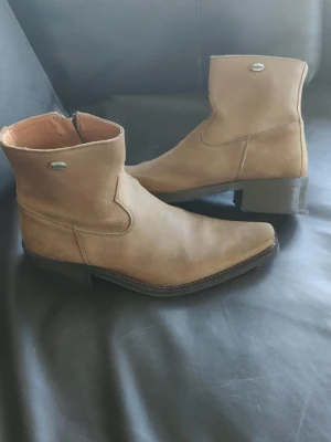 Oanvända Cowboy Boots i mocka - Snygga cowboy boots i beige mocka med spetsig tå och låg klack. Helt OANVÄNDA!!!Dragkedja på insidan för enkel påtagning och dekorativ söm längs skaftet. Perfekta för dig som vill ha en klassisk westernstil. Märke Dockers och para "provade". Blev aldrig av att jag använde dem pga jag bytte mode och stil. Hoppas "cowboy" kille/man kan få nytta av dem.