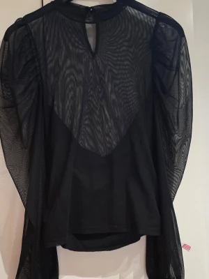 Svart meshblus med puffärmar - Trendig svart blus i mesh med långa puffärmar och rund halsringning. Blusen har transparent tyg på ärmar och överdel, samt en liten knapp i nacken. Perfekt för dig som vill sticka ut med en edgy och modern look.