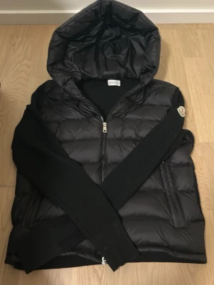 Svart dunjacka med huva från Moncler - Snygg svart dunjacka från Moncler med stickade ärmar och stor huva. Jackan har dragkedja framtill, två fickor och Moncler-logga på ena ärmen. Perfekt för kalla vinterdagar och riktigt trendig look. Pris kan diskuteras vid snabbt affär