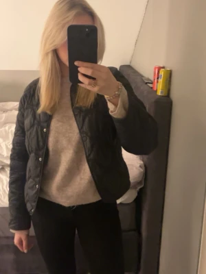 Svart quiltad jacka med knappar - Stilren svart quiltad jacka med tryckknappar framtill. Jackan har en klassisk passform och är lätt vadderad, perfekt för lager på lager. Passar till både jeans och kjol för en avslappnad look.