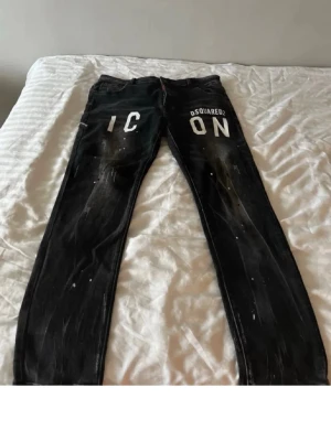 Svarta Dsquared2 ICON jeans - Snygga svarta jeans från Dsquared2 med vit 'ICON'-text framtill. Slim fit-modell med coola slitningar och stänkdetaljer för en trendig streetstyle-look. Perfekta för dig som vill sticka ut med en unik design och exklusiv känsla.