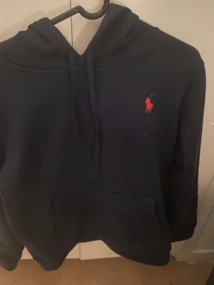 Mörkblå hoodie från Ralph Lauren - Mörkblå hoodie från Ralph Lauren med klassisk röd broderad logga på bröstet. Hoodien har huva med dragsko och en stor magficka framtill. Perfekt för en avslappnad och stilren look.
