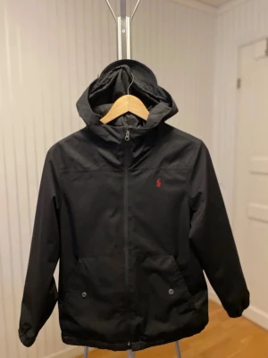 Svart vindjacka från Polo Ralph Lauren - Svart vindjacka med huva från Polo Ralph Lauren. Jackan har dragkedja framtill, två stora fickor med knappar och en liten röd logga på bröstet. Perfekt för blåsiga dagar och enkel att matcha med allt. Klassisk och stilren design.