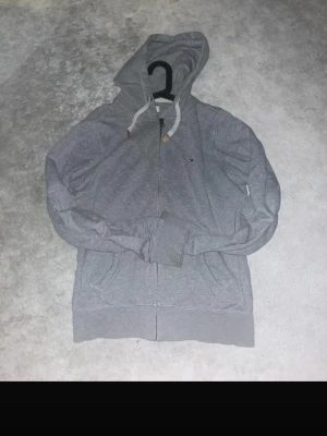 Grå Hilfiger Denim hoodie med dragkedja - Snygg grå hoodie från Hilfiger Denim med dragkedja framtill och två fickor. Klassisk huva med vita snören och diskret logga på bröstet. Tillverkad i mjukt bomullsmaterial som är skönt att ha på sig. Perfekt för en avslappnad och stilren look.