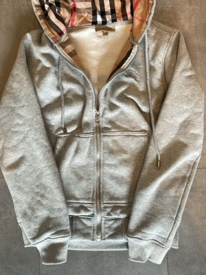 Grå hoodie från Burberry med dragkedja - Snygg grå hoodie från Burberry med klassiskt rutigt foder i huvan. Tröjan har dragkedja, två fickor fram och ribbade muddar. Materialet är mjukt och bekvämt, perfekt för en chill look. Ikoniskt Burberry-mönster på insidan av huvan ger extra stil.