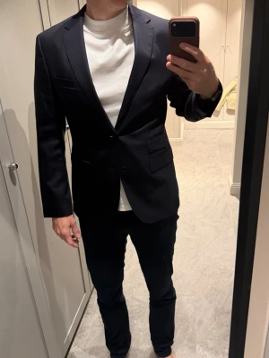 Mörkblå kavaj från BOSS - Stilren mörkblå kavaj från BOSS med klassisk passform och travel edition som inte blir skrynklig som vanliga kavajer. Kavajen har två framfickor med lock, bröstficka och knappar på ärmsluten. Materialet är slätt och ger ett elegant intryck. Perfekt för dig som vill ha en snygg och tidlös look.